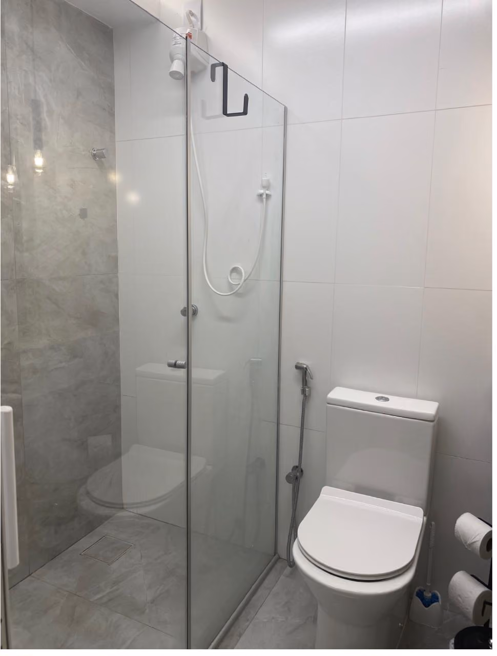 Cozinha compacta e moderna com bancada preta, pia e fogão de indução, prateleiras com eletrodomésticos e planta, e área de estar ao fundo com sofá preto, mesa branca e janela com cortina branca.