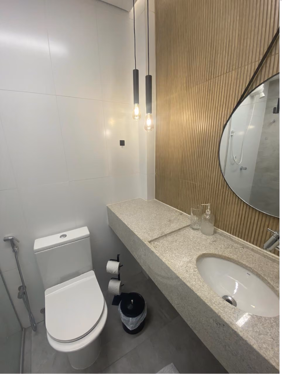 Sala de reunião ou espera com sofá preto de madeira à esquerda, parede azul ao fundo, fileira de cinco cadeiras claras à direita junto a uma bancada branca sob uma janela grande com persiana fechada, e uma TV de tela plana montada na parede próxima a uma porta branca no centro da parede ao fundo.