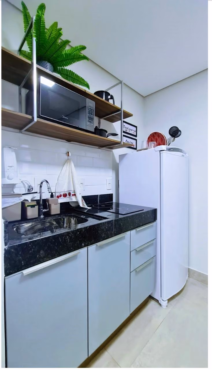 Quarto moderno com cama de solteiro branca com cobertor bege e roupas dobradas, TV preta sobre bancada branca, cadeira branca de escritório e duas pinturas abstratas com folhas e formas geométricas em tons terrosos na parede cinza clara.