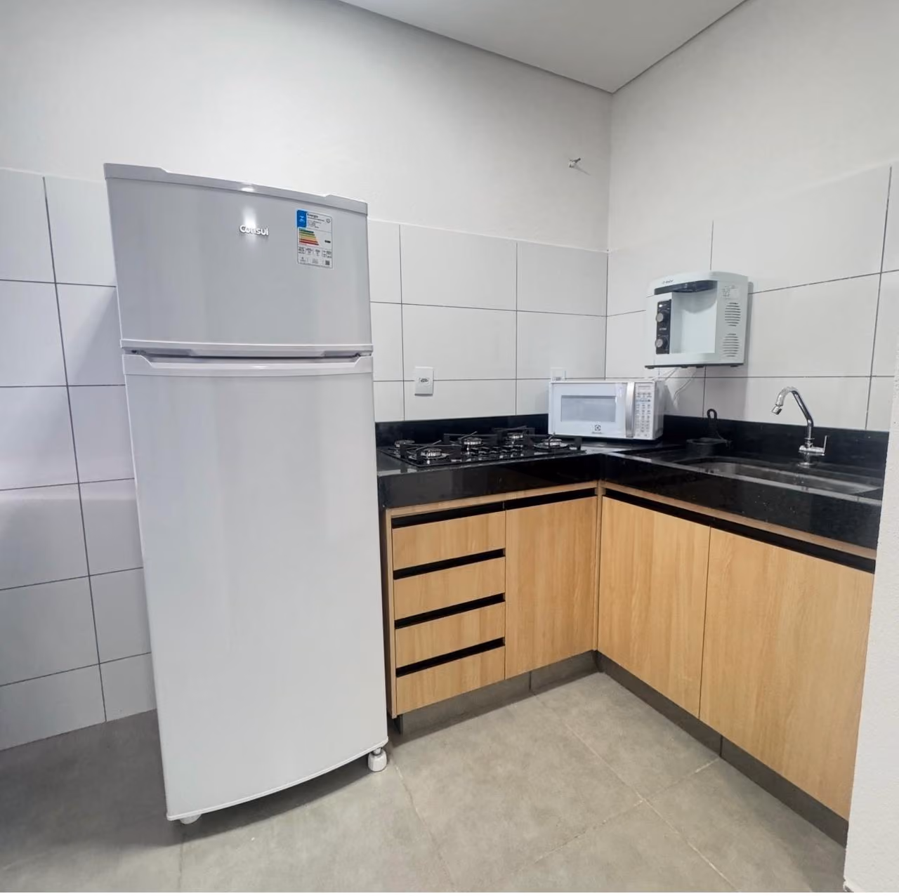Cozinha pequena com bancada de granito clara que tem cafeteira, fogão elétrico de duas bocas, pia de inox com escorredor, micro-ondas e frigobar branco, parede com grande espelho e porta de madeira com aviso de proibido fumar.