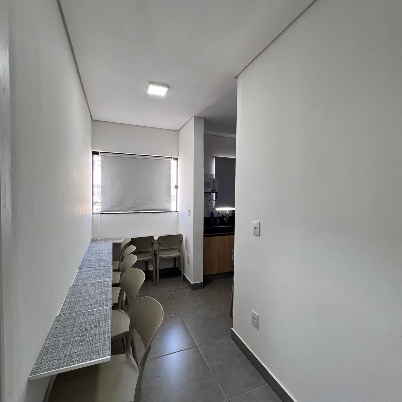 Cozinha pequena com bancada de granito clara que tem cafeteira, fogão elétrico de duas bocas, pia de inox com escorredor, micro-ondas e frigobar branco, parede com grande espelho e porta de madeira com aviso de proibido fumar.