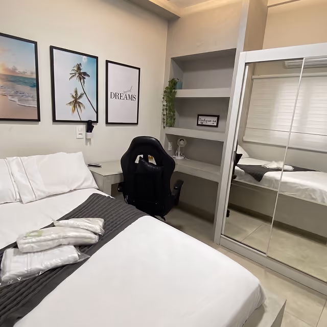 Quarto moderno com cama de casal e roupa de cama branca e cinza, cadeira de escritório preta ao lado de mesa de cabeceira, três quadros na parede com imagens de praia e texto, prateleiras embutidas com planta e itens decorativos, e guarda-roupa com portas de espelho refletindo o ambiente.